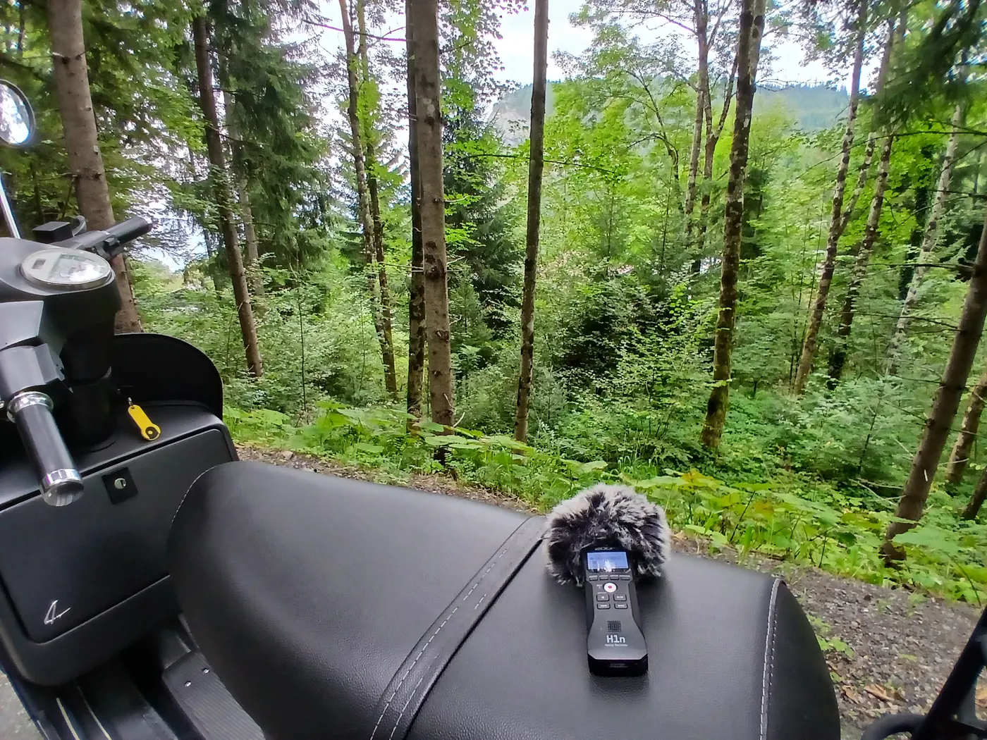 A recorder in a furry windshield resting on a moped seat in the middle of a forest track