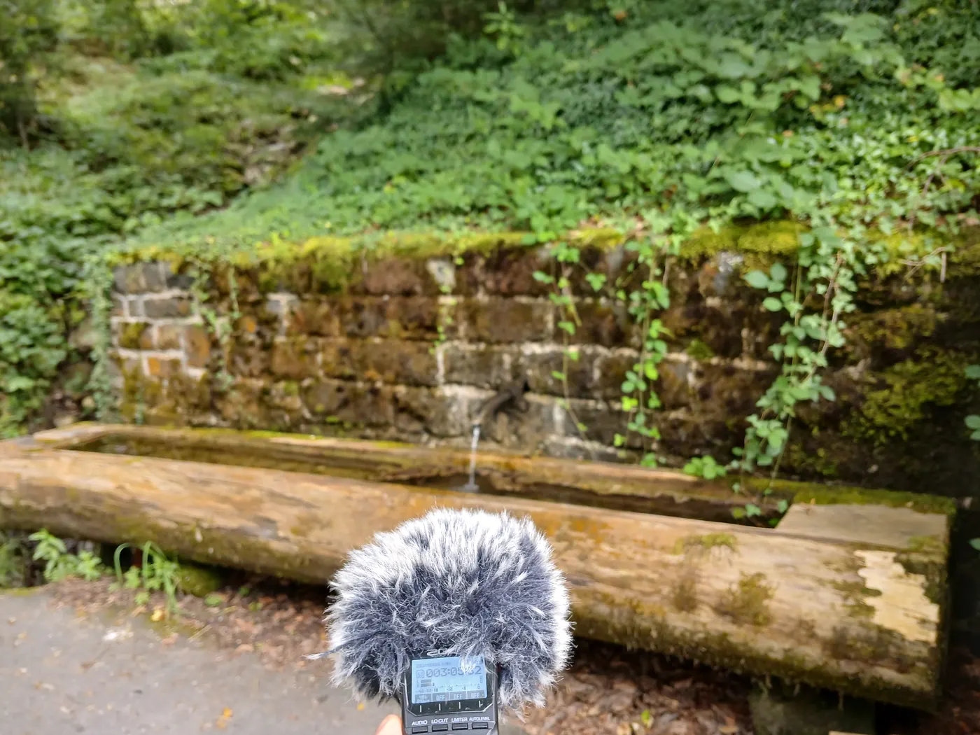 A recorder set up in front of an old stone water trough with a thin jet of water running into it