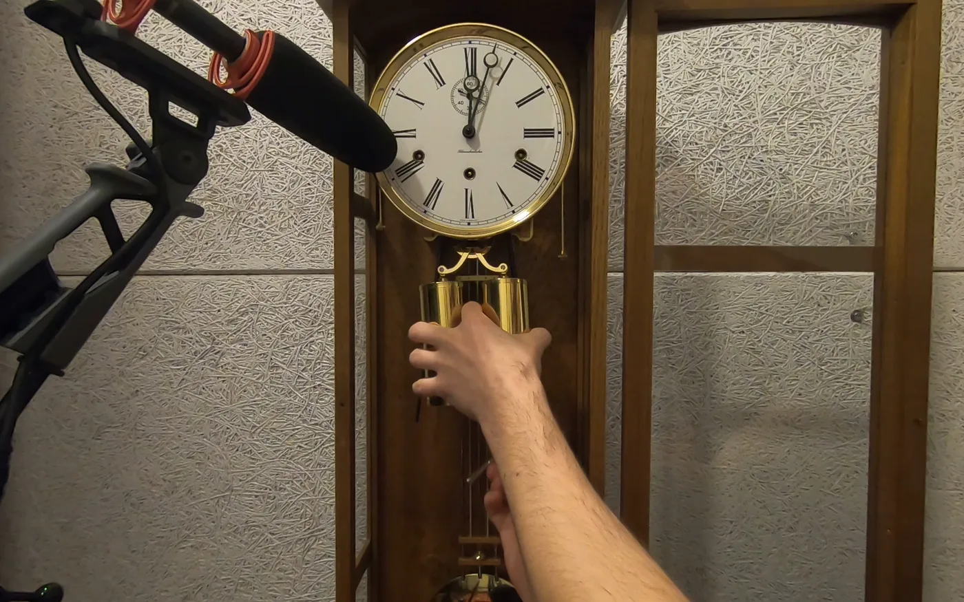 A shotgun microphone on a stand pointed into the open case of a Westminster chime clock, a hand damping the pendulum