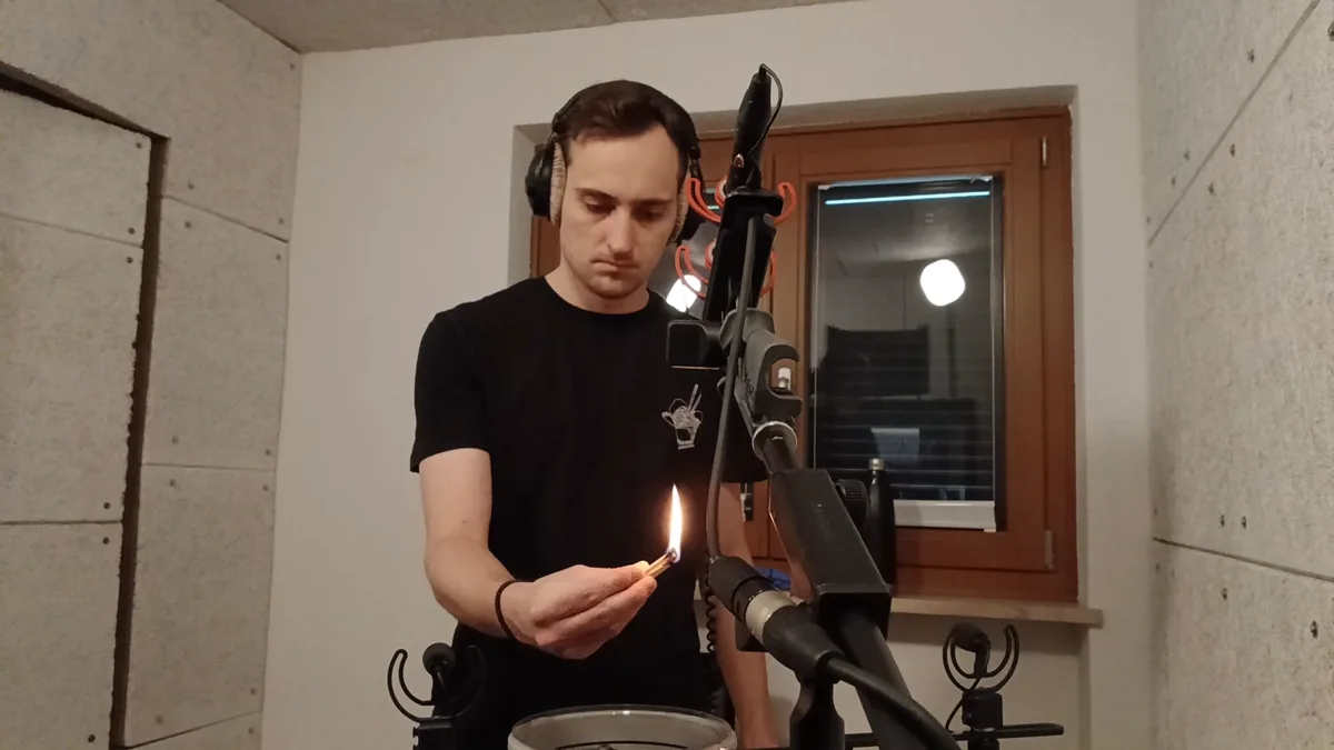 A man in headphones holding a lit lighter close to a microphone in the recording room, a second microphone over a cymbal in front of him